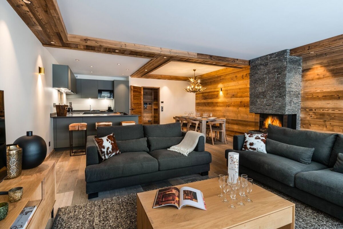 A cozy open-plan chalet living area with wood walls, stone fireplace, dark sofas, dining table, and modern kitchen. Rustic yet elegant design.