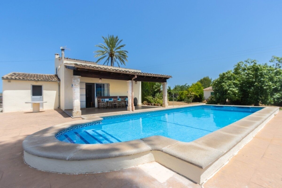 A sunny villa with a blue swimming pool, covered patio, and a palm tree under a clear sky, suggesting a relaxed vacation setting.