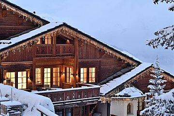 A beautiful, snow-laden wooden chalet illuminated by festive icicle lights and warm window glows, set against a winter mountain scene.