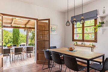A bright indoor dining area with a modern wooden table opens through French doors to an outdoor patio with another dining set and lush garden views.