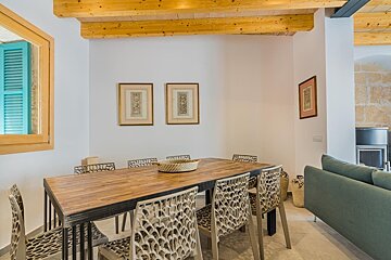 A cozy dining room featuring a long wooden table, modern chairs, exposed beams, and white walls with artwork. A blue-shutter window and a hint of a living area are visible.