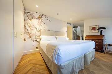 A serene bedroom with a large bed, cherry blossom wall art, wooden herringbone floor, and an antique desk. Stairs lead up in the background.