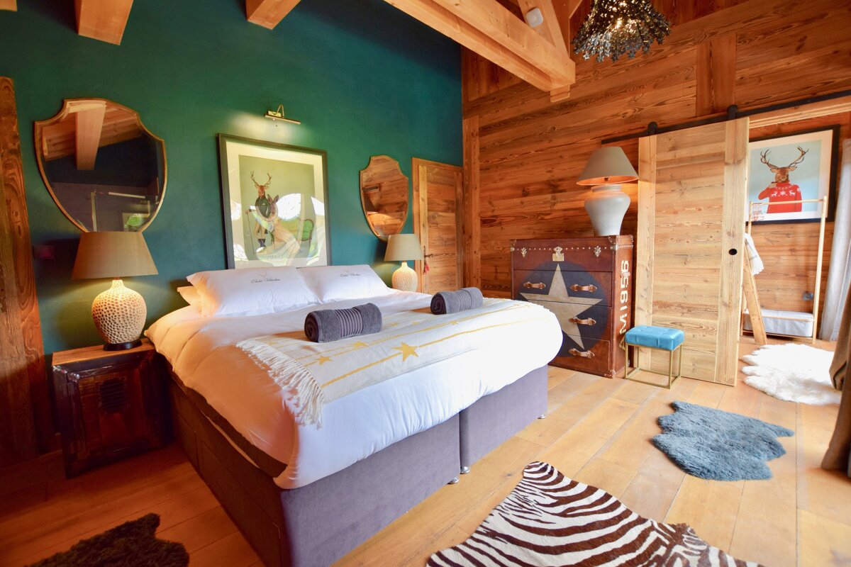 A cozy, rustic bedroom with a large bed, deep green accent wall, and warm wooden panels. Features include deer artwork, a vintage chest, and animal print rugs.