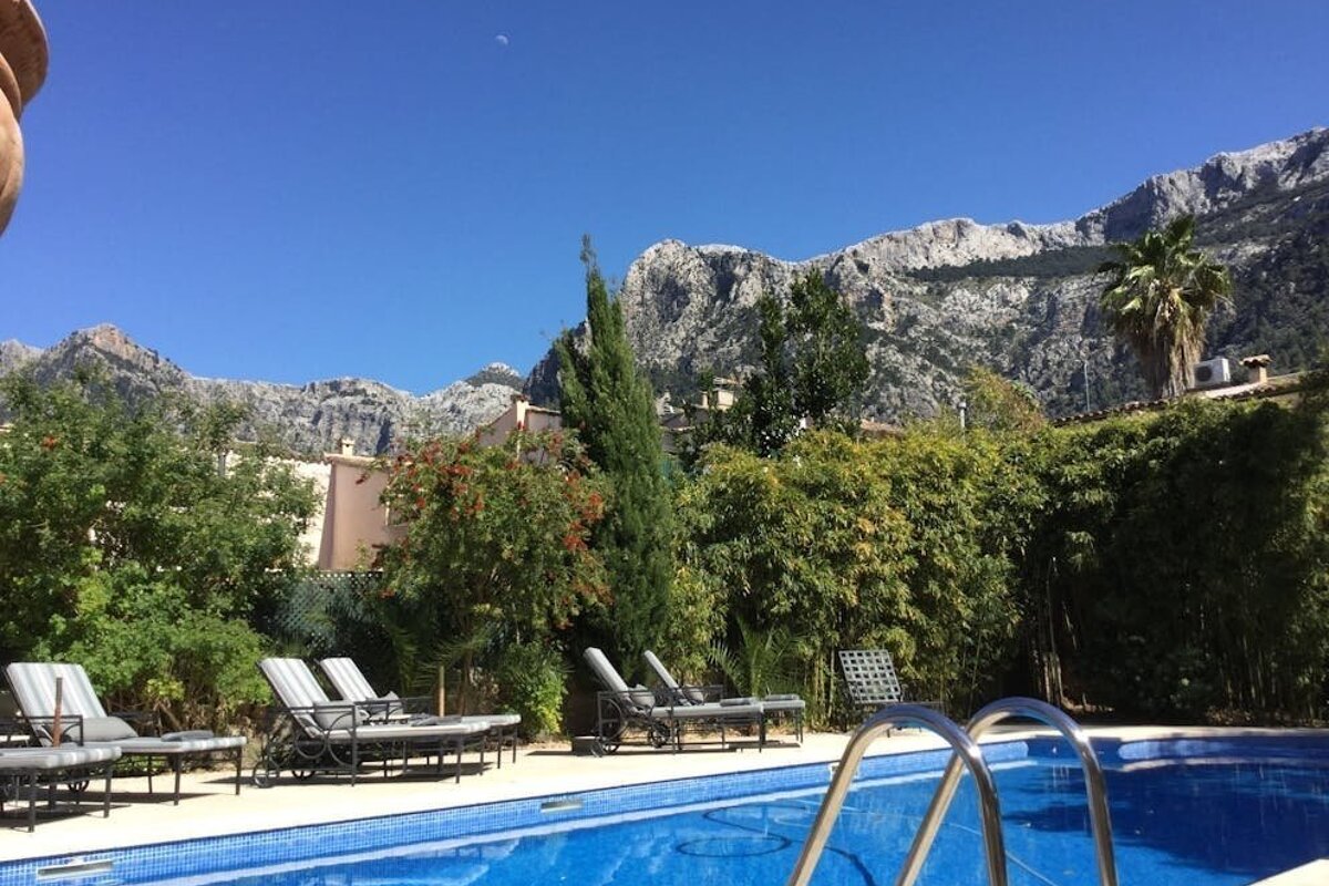 A blue tiled pool with lounge chairs, lush trees, and rocky mountains under a clear sky with a visible crescent moon.