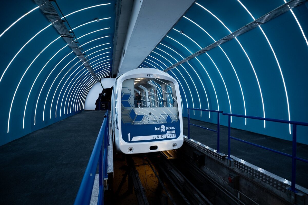 A sleek white funicular train sits in a futuristic blue tunnel station, illuminated by striking curved white lights. 
