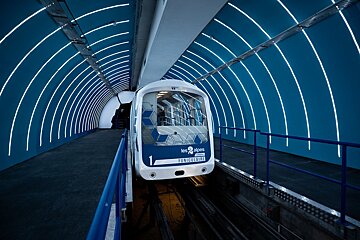 A sleek white funicular train sits in a futuristic blue tunnel station, illuminated by striking curved white lights. 