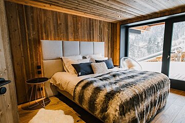 Cozy chalet bedroom with warm wood walls, a plush bed, and a large window framing a stunning snowy mountain view.