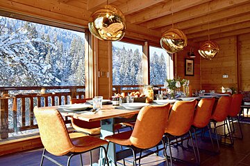 A cozy wooden dining room with a long table, orange chairs, and golden lights, overlooking a snowy forest through large windows.