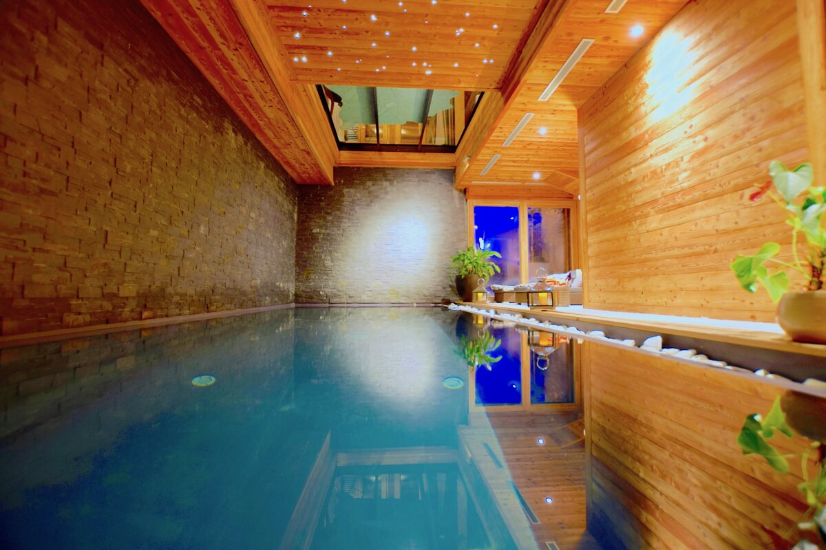An inviting indoor pool with warm wood paneling, stone walls, a starry ceiling, and blue reflections from windows, creating a serene spa atmosphere.