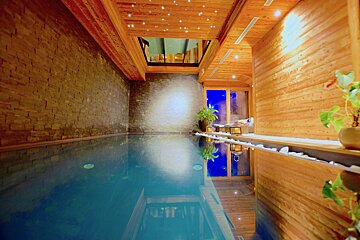 An inviting indoor pool with warm wood paneling, stone walls, a starry ceiling, and blue reflections from windows, creating a serene spa atmosphere.