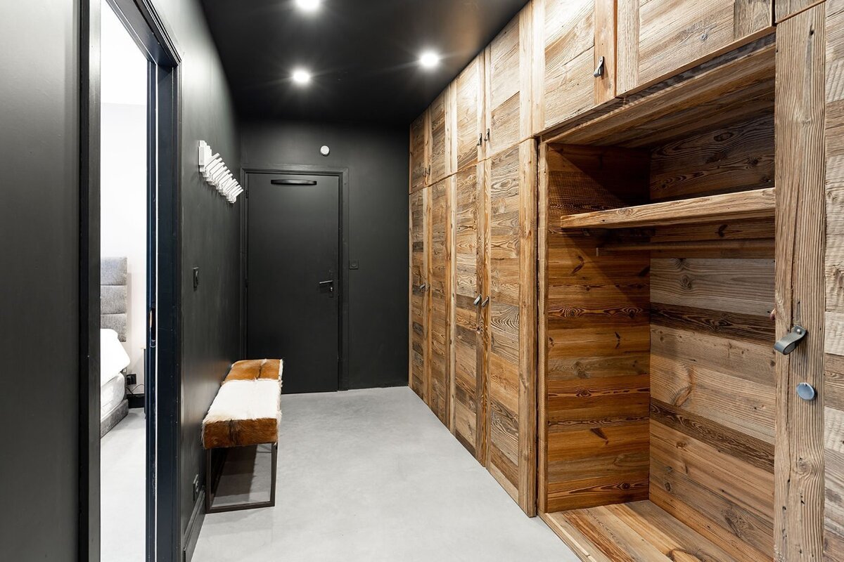 A modern hallway features dark walls and ceiling with recessed lights, contrasting with a long wall of rustic wooden built-in wardrobes. A fur-topped bench sits opposite dark doors.