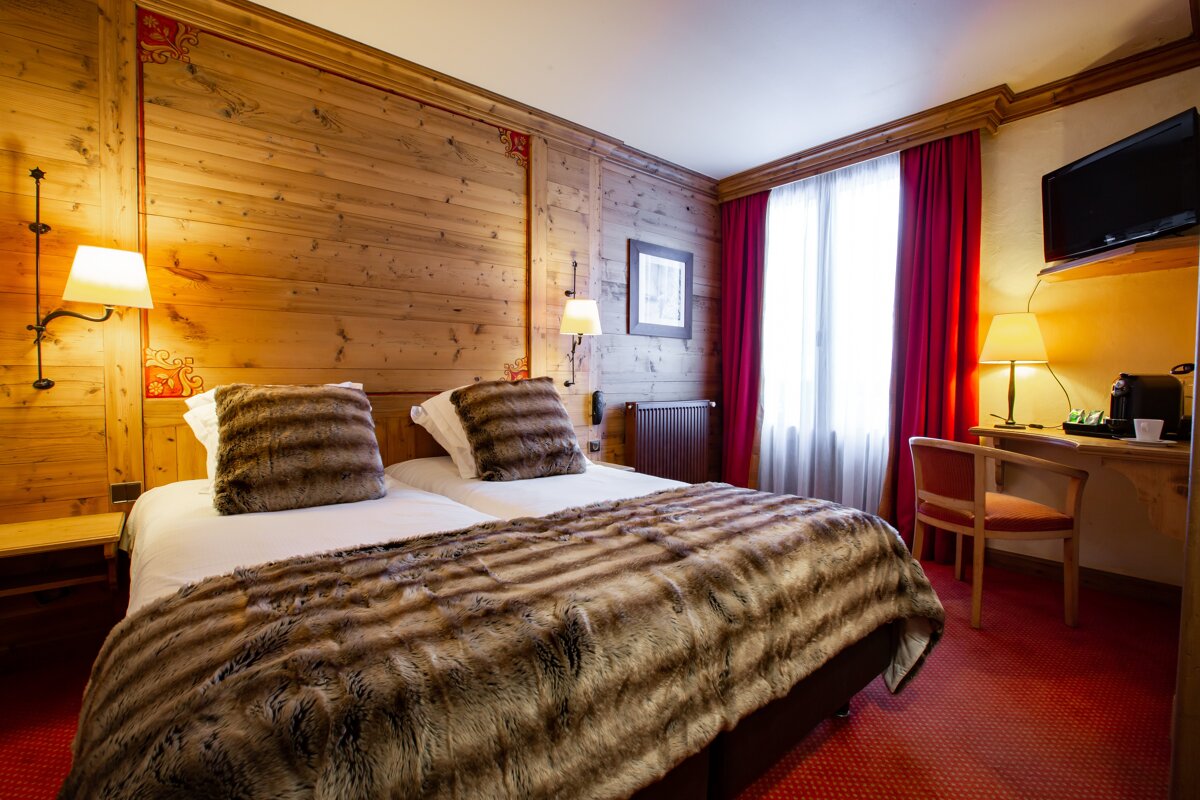 Cozy, chalet-style bedroom with wood-paneled walls, twin beds with furry throws, red curtains & carpet, and a desk with a TV.