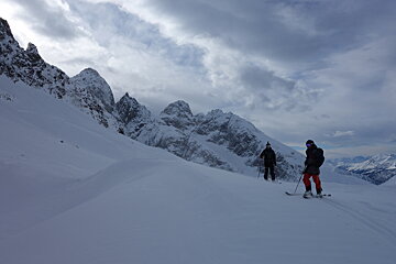Two people skiing down a snow covered slope with mountains in the background