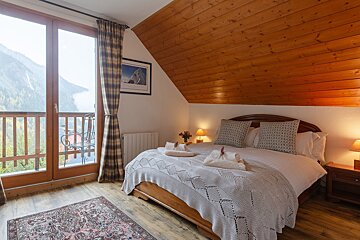 A cozy bedroom features a wooden bed, sloped ceiling, and a balcony opening to expansive mountain and valley views. Plaid curtains and a patterned rug complete the warm space.