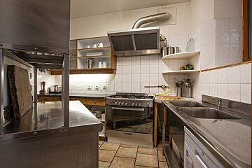 A functional kitchen with industrial-style stainless steel counters, a gas stove, double sink, white tiled walls, and open shelving.