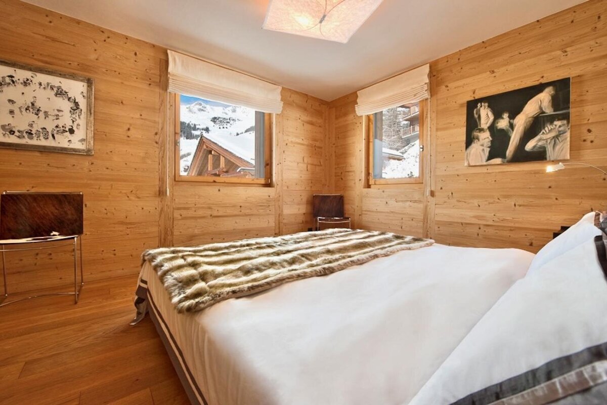 Cozy wooden bedroom with a large bed, fur throw, and two windows offering snowy mountain views. Art adorns the pine-paneled walls.