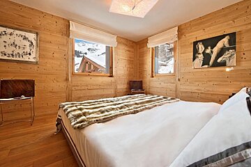 Cozy wooden bedroom with a large bed, fur throw, and two windows offering snowy mountain views. Art adorns the pine-paneled walls.