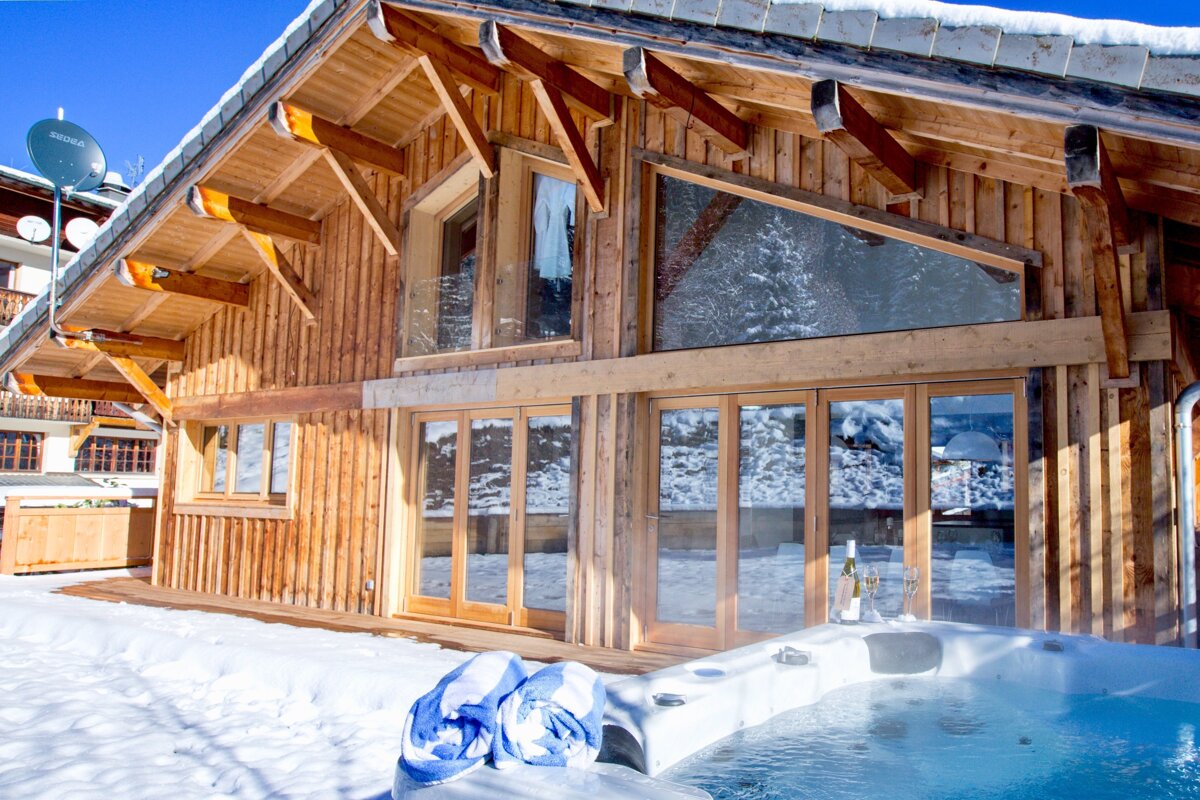 A rustic wooden chalet with large windows stands in a snowy landscape. An inviting hot tub, with towels and drinks, sits outside under a clear blue sky.