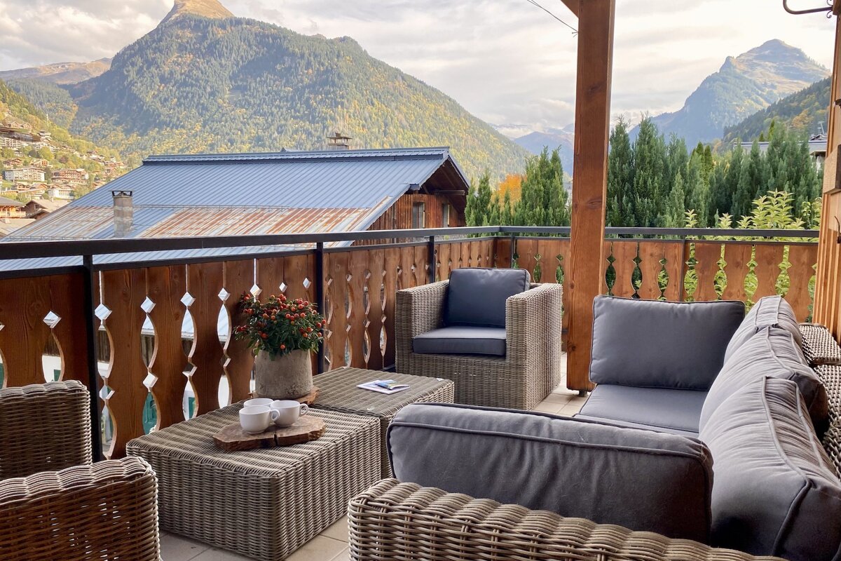 A cozy outdoor balcony with wicker seating and a stunning view of a mountain village and forested alpine peaks, perfect for relaxation.