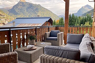 A cozy outdoor balcony with wicker seating and a stunning view of a mountain village and forested alpine peaks, perfect for relaxation.