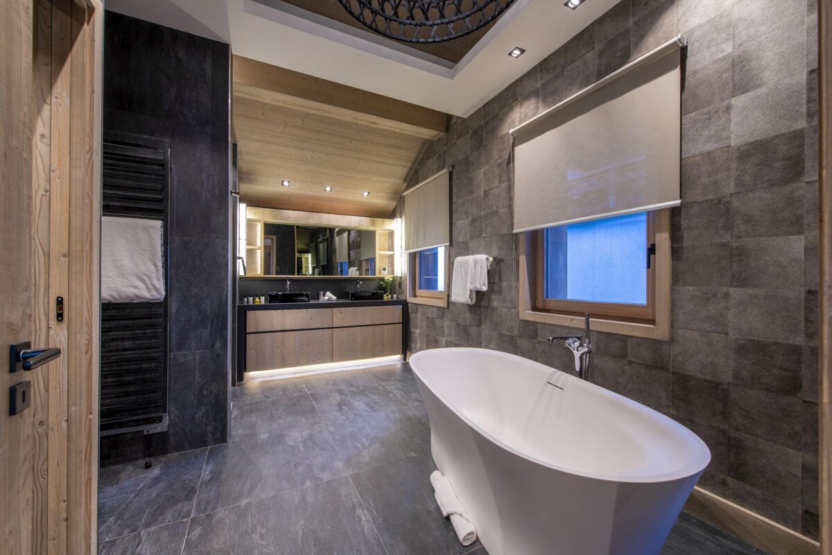 A luxurious modern bathroom features a white freestanding tub, dark slate-like tiles, a double wooden vanity with strip lighting, and warm wood accents.