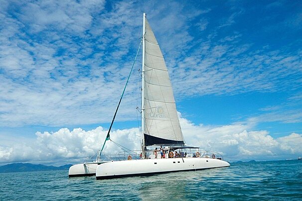 A white catamaran with a sail up cruises on blue water under a partly cloudy sky, carrying people and with distant land visible.