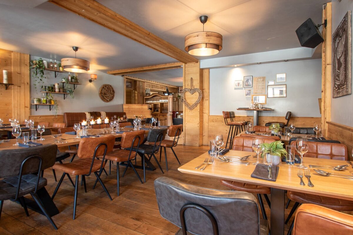 Cozy, rustic restaurant interior with warm lighting, wooden walls, beams, and floors. Features set tables, leather chairs, banquettes, plants, and framed art.