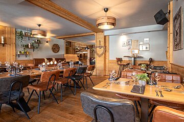 Cozy, rustic restaurant interior with warm lighting, wooden walls, beams, and floors. Features set tables, leather chairs, banquettes, plants, and framed art.
