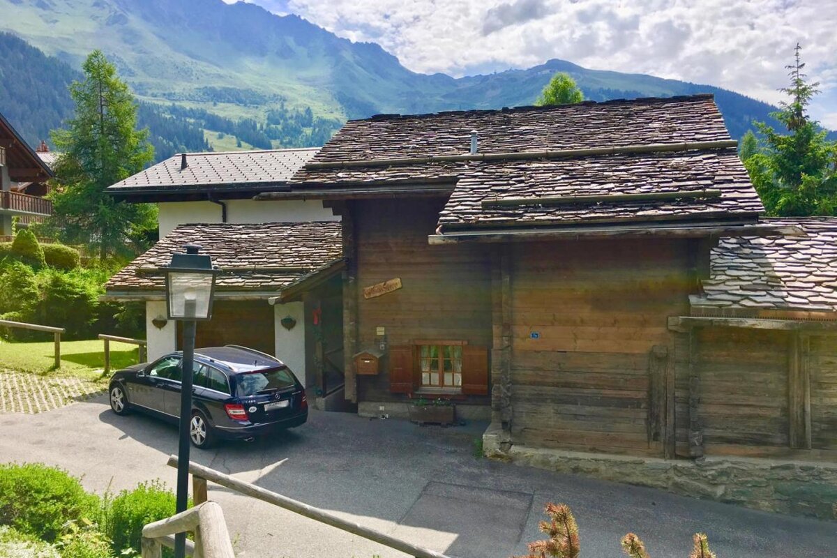 A car is parked in front of a wooden house with a mountain in the background