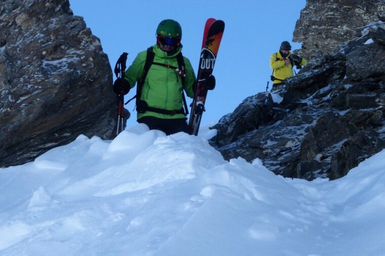 A man in a green jacket is carrying a pair of skis that say just
