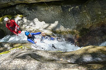 Two people in wetsuits and helmets are canyoning through a narrow, rocky gorge with rushing white water. One in red stands by, while another in blue navigates the current.