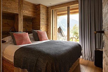 A cozy, rustic bedroom with wood-paneled walls, a bed with colorful pillows, and a sliding glass door revealing a scenic mountain view.