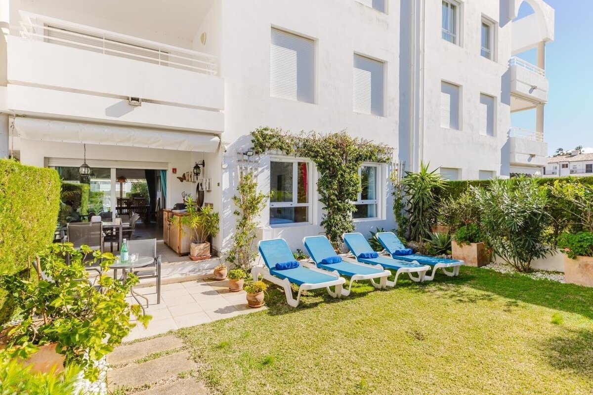 A bright white building with a shaded patio, outdoor dining area, and a green lawn with blue lounge chairs. Lush plants and bushes complete the sunny scene.