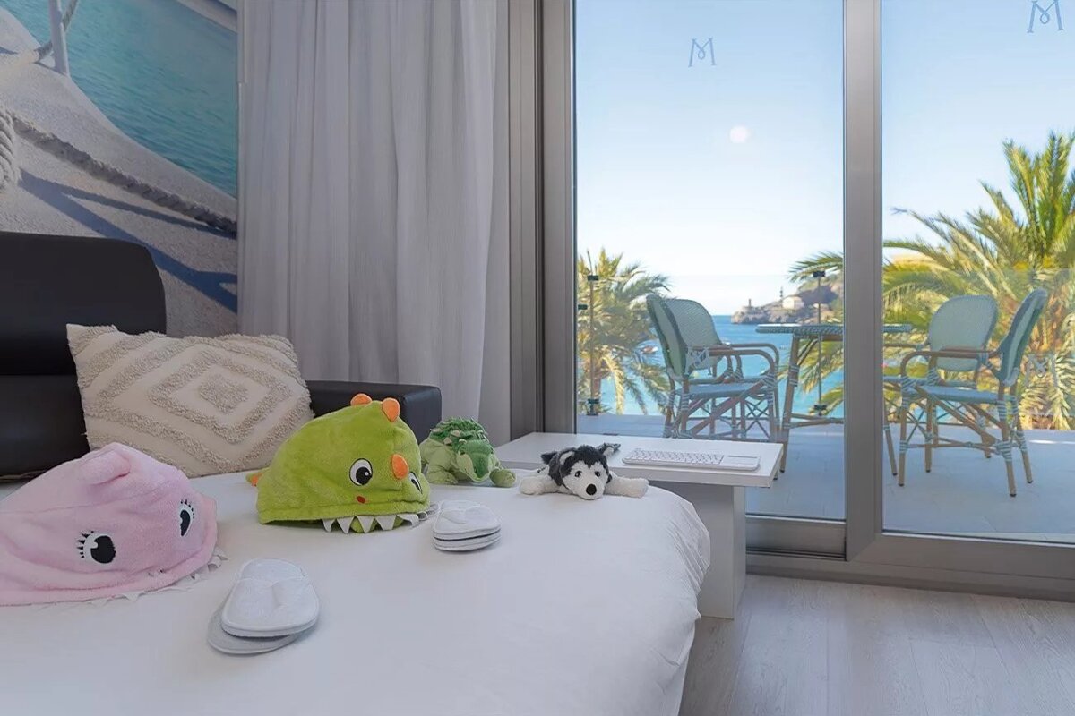 A bright room with stuffed animals and slippers on the bed, opening to a balcony with rattan chairs, palm trees, and a stunning sea view with a lighthouse.