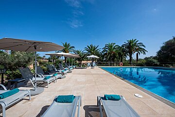 Sunny resort pool with rows of loungers, teal towels, umbrellas, and palm trees under a clear blue sky.