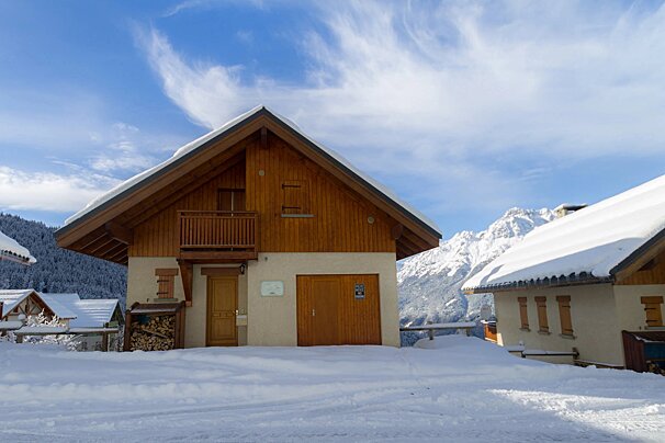 A wooden chalet stands in deep snow, backed by snow-capped mountains under a clear blue sky with wispy clouds.