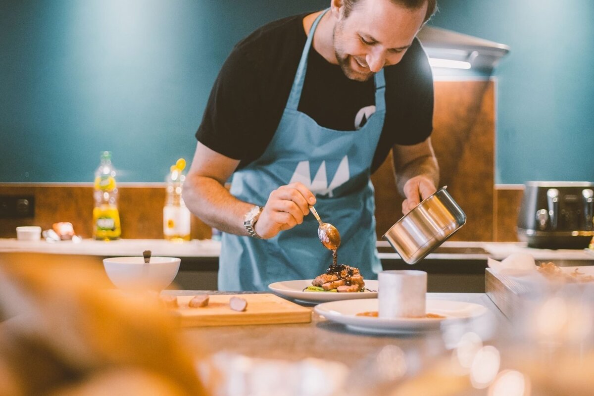 A smiling man in a blue apron meticulously garnishes a plated dish with sauce in a modern kitchen.