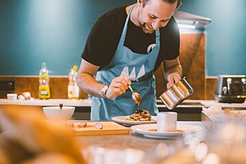A smiling man in a blue apron meticulously garnishes a plated dish with sauce in a modern kitchen.