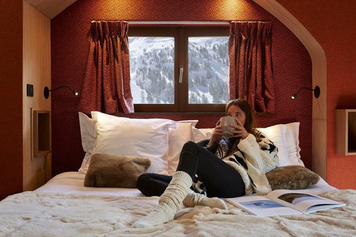 A woman sits on a bed reading a book and drinking from a mug