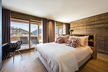 A cozy chalet bedroom with wooden walls and floor, a large bed with colorful pillows, and a balcony offering a picturesque view of snowy mountains.