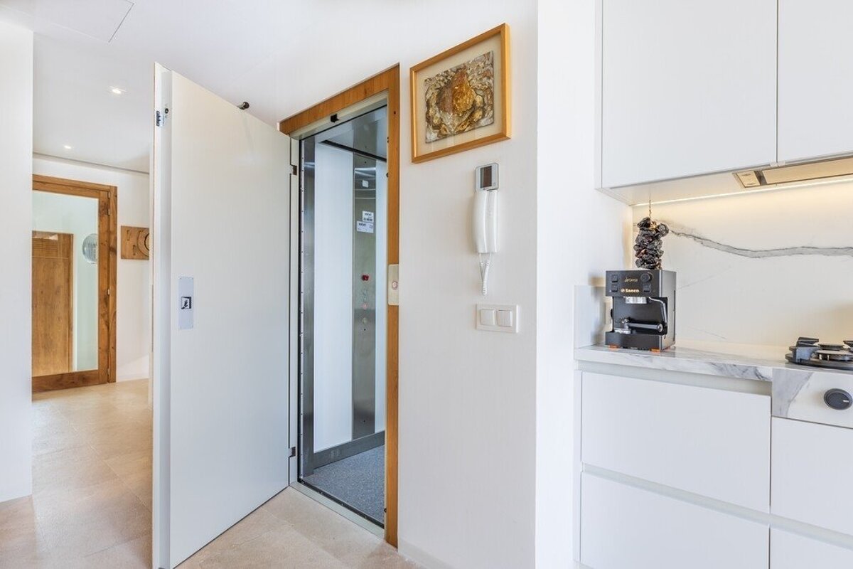 A modern interior featuring an open elevator next to a white door, an intercom, framed art, and a sleek kitchen with a coffee machine.