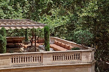Elegant rooftop terrace with shaded brown seating, potted plants, and ornate stone railing, set against a lush green forest backdrop.