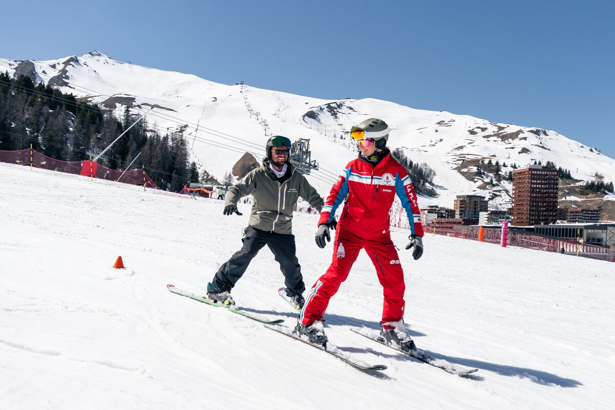 A ski instructor in red teaches a student in green on a sunny snowy slope. Mountains, ski lifts, and resort buildings are visible in the background.