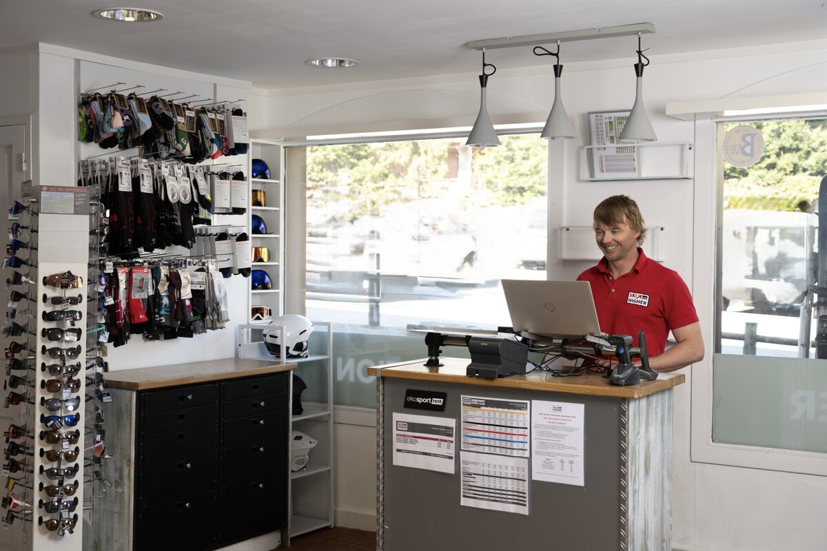 A smiling man in a red shirt works on a laptop behind a counter in a ski/sports shop displaying helmets, goggles, and accessories.