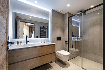 A modern bathroom features grey tiled walls, a wooden vanity with a lit mirror, a floating toilet, and a sleek glass-enclosed shower.