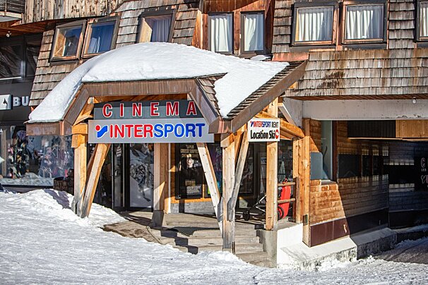 A snow-covered wooden building entrance houses a cinema and Intersport ski rental shop. Snowy ground leads up to the steps.