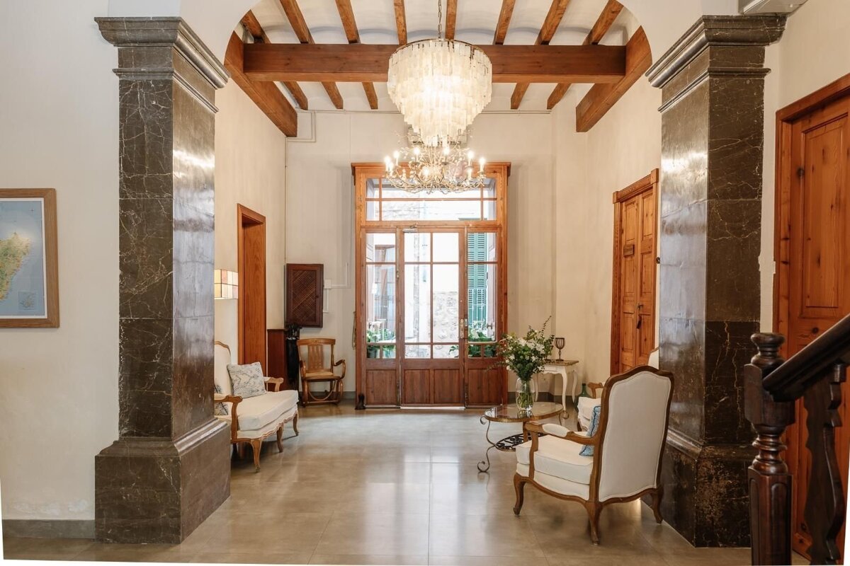A grand, elegant hallway with dark marble pillars, exposed wooden beams, and two chandeliers. French Provincial furniture and a large glass door adorn the space.