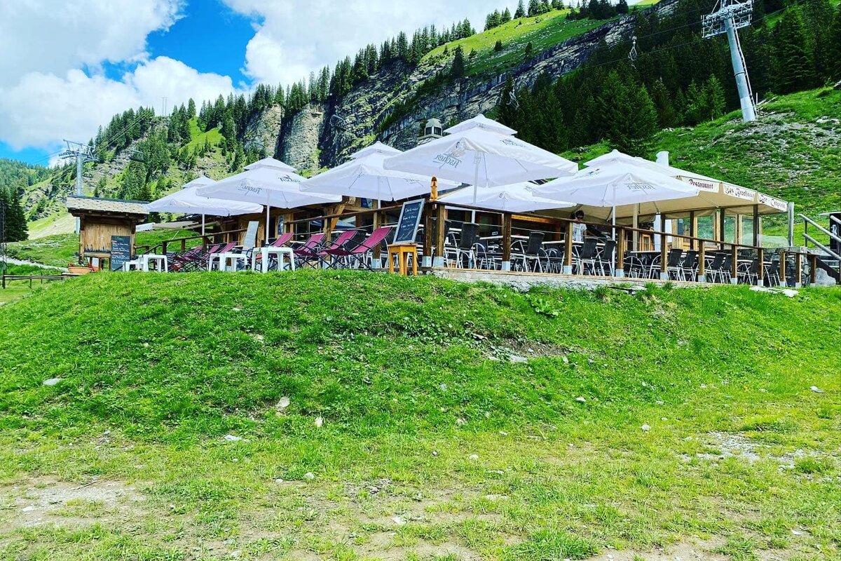 A mountain restaurant with a large outdoor patio shaded by white umbrellas, nestled on a grassy slope with pine trees, rocky cliffs, and a ski lift in the background.