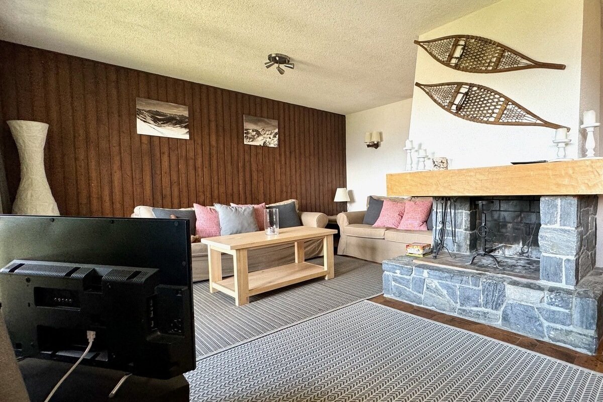 A cozy, rustic living room featuring wood-paneled walls, a stone fireplace with snowshoe decor, sofas, coffee table, and a TV.