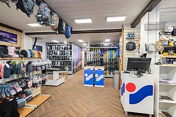 A brightly lit ski shop interior displays ski boots, skis, winter hats, gloves, and backpacks. It's a well-organized retail space for winter sports gear.